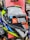 A close-up view of a motorcycle helmet is displayed prominently against a red background. The helmet features a mixture of bright and reflective colors, including neon yellow, blue, white, black, and silver. There is a large sticker on the visor with text reading 'Designed to Protect' and a website URL 'www.mthelmets.com'. A blue sticker indicating quality control pass (QC PASSED) is also affixed near the visor mechanism. The environment shows other helmets and equipment, indicating a store setting.