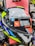 A close-up view of a motorcycle helmet is displayed prominently against a red background. The helmet features a mixture of bright and reflective colors, including neon yellow, blue, white, black, and silver. There is a large sticker on the visor with text reading 'Designed to Protect' and a website URL 'www.mthelmets.com'. A blue sticker indicating quality control pass (QC PASSED) is also affixed near the visor mechanism. The environment shows other helmets and equipment, indicating a store setting.