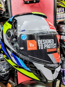A close-up view of a motorcycle helmet is displayed prominently against a red background. The helmet features a mixture of bright and reflective colors, including neon yellow, blue, white, black, and silver. There is a large sticker on the visor with text reading 'Designed to Protect' and a website URL 'www.mthelmets.com'. A blue sticker indicating quality control pass (QC PASSED) is also affixed near the visor mechanism. The environment shows other helmets and equipment, indicating a store setting.