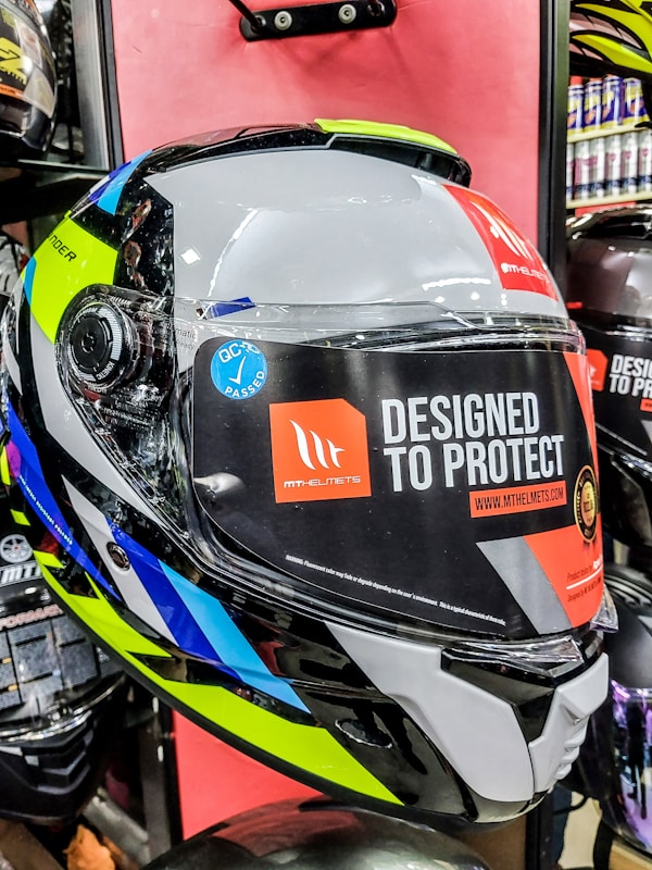 A close-up view of a motorcycle helmet is displayed prominently against a red background. The helmet features a mixture of bright and reflective colors, including neon yellow, blue, white, black, and silver. There is a large sticker on the visor with text reading 'Designed to Protect' and a website URL 'www.mthelmets.com'. A blue sticker indicating quality control pass (QC PASSED) is also affixed near the visor mechanism. The environment shows other helmets and equipment, indicating a store setting.