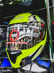 A motorcycle helmet is displayed with a bright yellow and camouflage design. The face shield is labeled with text indicating it is a scratch-resistant visor. The helmet is placed on a shelf with other helmets visible behind it.