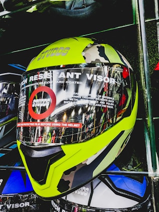 A motorcycle helmet is displayed with a bright yellow and camouflage design. The face shield is labeled with text indicating it is a scratch-resistant visor. The helmet is placed on a shelf with other helmets visible behind it.
