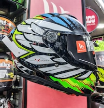 A brightly colored motorcycle helmet is displayed, featuring a bold, intricate design with patterns in black, white, blue, green, and red. It is positioned in a retail setting, surrounded by other helmets. The visor has a sticker with branding and text indicating the design concept.