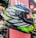 A brightly colored motorcycle helmet is displayed, featuring a bold, intricate design with patterns in black, white, blue, green, and red. It is positioned in a retail setting, surrounded by other helmets. The visor has a sticker with branding and text indicating the design concept.