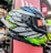 A brightly colored motorcycle helmet is displayed, featuring a bold, intricate design with patterns in black, white, blue, green, and red. It is positioned in a retail setting, surrounded by other helmets. The visor has a sticker with branding and text indicating the design concept.