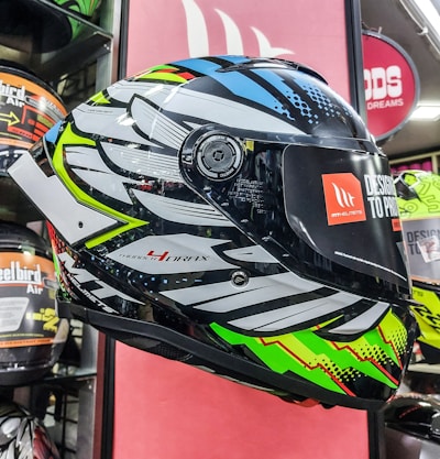 A brightly colored motorcycle helmet is displayed, featuring a bold, intricate design with patterns in black, white, blue, green, and red. It is positioned in a retail setting, surrounded by other helmets. The visor has a sticker with branding and text indicating the design concept.