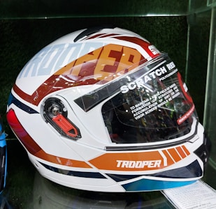 A detailed view of a motorcycle helmet with a glossy design that includes bold text and geometric patterns. The helmet features words like 'TROOPER' and 'SCRATCH RESISTANT' across the surface, with technical details and a visible visor mechanism.
