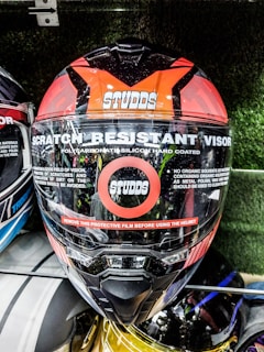 A motorcycle helmet prominently displays features such as a scratch-resistant visor and polycarbonate silicon hard coating. The brand name Studds is visible on the front. A red stripe warns users to remove the protective film before use. The helmet is among others in a display setting.