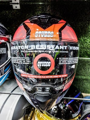 A motorcycle helmet prominently displays features such as a scratch-resistant visor and polycarbonate silicon hard coating. The brand name Studds is visible on the front. A red stripe warns users to remove the protective film before use. The helmet is among others in a display setting.