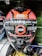 A motorcycle helmet prominently displays features such as a scratch-resistant visor and polycarbonate silicon hard coating. The brand name Studds is visible on the front. A red stripe warns users to remove the protective film before use. The helmet is among others in a display setting.