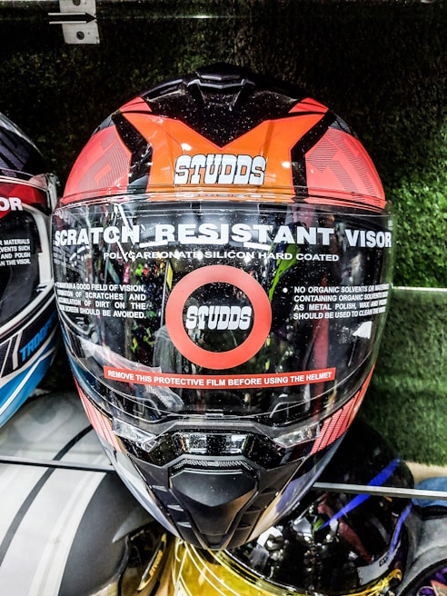 A motorcycle helmet prominently displays features such as a scratch-resistant visor and polycarbonate silicon hard coating. The brand name Studds is visible on the front. A red stripe warns users to remove the protective film before use. The helmet is among others in a display setting.