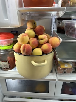 A refrigerator shelf with a pot filled with fresh peaches. The pot is surrounded by other containers and jars inside the refrigerator.