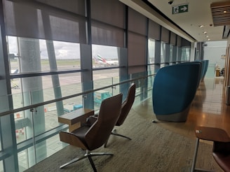 A modern airport lounge with comfortable seating positioned along large windows providing a view of the tarmac and parked aircraft. The area is well-lit with natural light filtering through partially drawn blinds, and features sleek, contemporary furniture including cushioned chairs and small tables. An exit sign is visible on the ceiling.