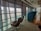 A modern airport lounge with comfortable seating positioned along large windows providing a view of the tarmac and parked aircraft. The area is well-lit with natural light filtering through partially drawn blinds, and features sleek, contemporary furniture including cushioned chairs and small tables. An exit sign is visible on the ceiling.