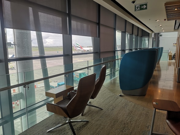 A modern airport lounge with comfortable seating positioned along large windows providing a view of the tarmac and parked aircraft. The area is well-lit with natural light filtering through partially drawn blinds, and features sleek, contemporary furniture including cushioned chairs and small tables. An exit sign is visible on the ceiling.