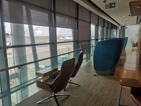 A modern airport lounge with comfortable seating positioned along large windows providing a view of the tarmac and parked aircraft. The area is well-lit with natural light filtering through partially drawn blinds, and features sleek, contemporary furniture including cushioned chairs and small tables. An exit sign is visible on the ceiling.