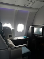 A luxurious airplane cabin features a large, comfortable seat with a plush pillow. Adjacent to the seat is a personal entertainment screen and a side table with items including a magazine. The cabin is dimly lit with a soft ambiance from overhead lighting, and two windows allowing some muted daylight.