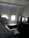 A luxurious airplane cabin features a large, comfortable seat with a plush pillow. Adjacent to the seat is a personal entertainment screen and a side table with items including a magazine. The cabin is dimly lit with a soft ambiance from overhead lighting, and two windows allowing some muted daylight.