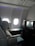 A luxurious airplane cabin features a large, comfortable seat with a plush pillow. Adjacent to the seat is a personal entertainment screen and a side table with items including a magazine. The cabin is dimly lit with a soft ambiance from overhead lighting, and two windows allowing some muted daylight.