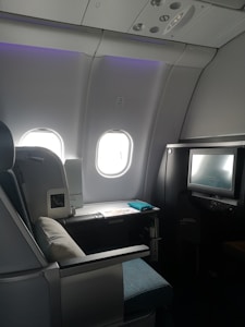 A luxurious airplane cabin features a large, comfortable seat with a plush pillow. Adjacent to the seat is a personal entertainment screen and a side table with items including a magazine. The cabin is dimly lit with a soft ambiance from overhead lighting, and two windows allowing some muted daylight.