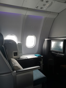 A luxurious airplane cabin features a large, comfortable seat with a plush pillow. Adjacent to the seat is a personal entertainment screen and a side table with items including a magazine. The cabin is dimly lit with a soft ambiance from overhead lighting, and two windows allowing some muted daylight.