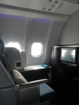 A luxurious airplane cabin features a large, comfortable seat with a plush pillow. Adjacent to the seat is a personal entertainment screen and a side table with items including a magazine. The cabin is dimly lit with a soft ambiance from overhead lighting, and two windows allowing some muted daylight.