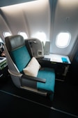 A luxurious airplane seat with a teal upholstery, complemented by a white pillow. It is situated next to a window section, revealing several airplane windows in the cabin. The compartment includes a tray table with a magazine, a small screen, and a control panel.