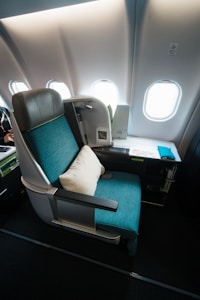 A luxurious airplane seat with a teal upholstery, complemented by a white pillow. It is situated next to a window section, revealing several airplane windows in the cabin. The compartment includes a tray table with a magazine, a small screen, and a control panel.