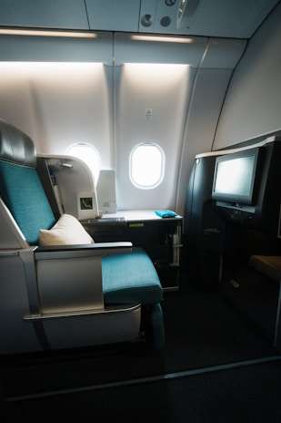 A luxurious airplane seat featuring a turquoise cushion and a white pillow is situated next to a window. The seat is part of a first-class or business-class section with ample space and a modern design. There is a television screen integrated into a partition in front of the seat, and a small storage area is present with various amenities. Natural light from the window illuminates the area, casting soft shadows across the plush fabric and sleek surfaces.