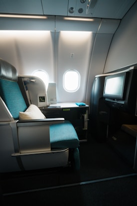 A luxurious airplane seat featuring a turquoise cushion and a white pillow is situated next to a window. The seat is part of a first-class or business-class section with ample space and a modern design. There is a television screen integrated into a partition in front of the seat, and a small storage area is present with various amenities. Natural light from the window illuminates the area, casting soft shadows across the plush fabric and sleek surfaces.