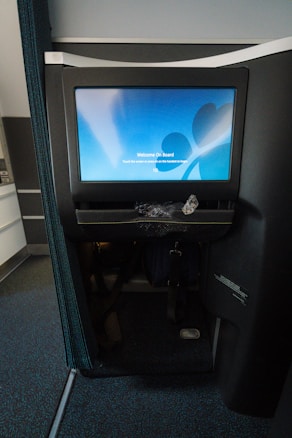 A personal screen display with a welcome message is installed in an airplane seat. The screen shows a blue welcome screen with a message to touch the screen or use the handset to begin. Below the screen, there is a netted pocket possibly containing headphones packaged in plastic. The surrounding area includes parts of the airplane seat and cabin.