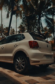 A white car is parked on a checkered pavement surrounded by tall palm trees. The vehicle is positioned at an angle, showcasing its rear and side, with the license plate partially visible. The background is filled with a blue sky and tropical foliage.