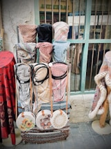 A display of traditional woven garments and accessories, including keffiyehs in various patterns and colors, hangs against a rustic background. Round woven bags with floral and intricate designs are neatly arranged below the garments. The setting appears to be a market or shop, characterized by its aged wall and weathered window frame.