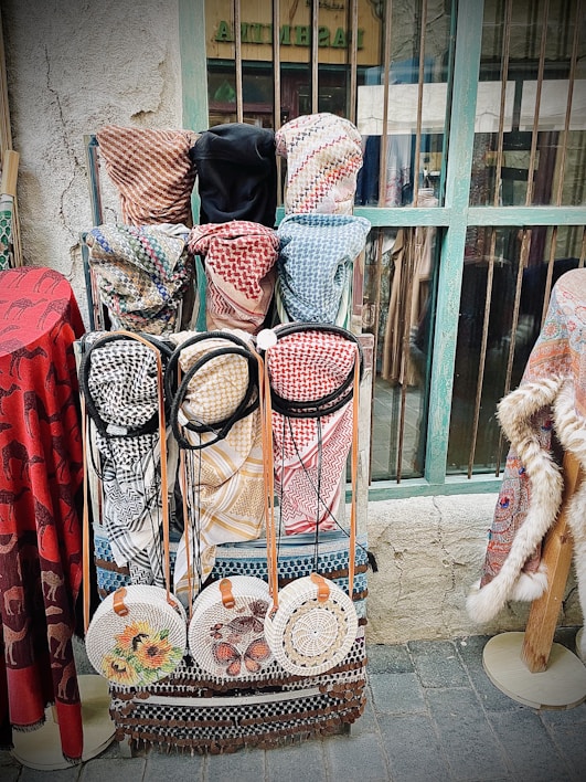 A display of traditional woven garments and accessories, including keffiyehs in various patterns and colors, hangs against a rustic background. Round woven bags with floral and intricate designs are neatly arranged below the garments. The setting appears to be a market or shop, characterized by its aged wall and weathered window frame.