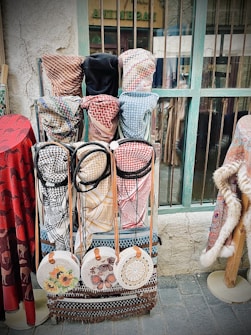 A display of traditional woven garments and accessories, including keffiyehs in various patterns and colors, hangs against a rustic background. Round woven bags with floral and intricate designs are neatly arranged below the garments. The setting appears to be a market or shop, characterized by its aged wall and weathered window frame.