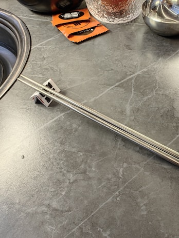 A gray marble-like countertop with a metal object resembling a handle or a strip, positioned along it. To the top left, part of a stainless steel bowl is visible. There are two orange packages with black and white labels, partially visible at the top. A clear glass with textured surface and a small metal bowl are present on the upper right corner.