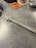 A gray marble-like countertop with a metal object resembling a handle or a strip, positioned along it. To the top left, part of a stainless steel bowl is visible. There are two orange packages with black and white labels, partially visible at the top. A clear glass with textured surface and a small metal bowl are present on the upper right corner.