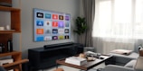A modern living room featuring a mounted flat-screen TV displaying various streaming service icons. The room includes a black entertainment console beneath the TV, a brown wooden coffee table with books and remote controls, a gray sofa, and a tall green plant near a large window with sheer white curtains.