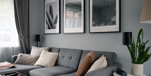A modern living room features a stylish gray sectional sofa adorned with assorted cushions in neutral tones. Above the sofa, three large black-and-white framed photographs hang on the wall. Natural light filters in through a window covered with semi-transparent curtains. A green potted plant sits on a table on the right side, adding a touch of nature to the space.