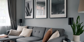 A modern living room features a stylish gray sectional sofa adorned with assorted cushions in neutral tones. Above the sofa, three large black-and-white framed photographs hang on the wall. Natural light filters in through a window covered with semi-transparent curtains. A green potted plant sits on a table on the right side, adding a touch of nature to the space.