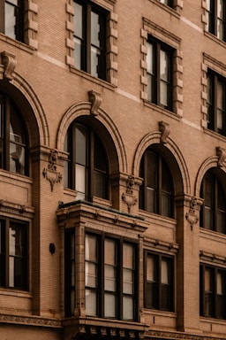 An architectural close-up of a historic building facade featuring ornate details such as decorative moldings, brickwork, and arched windows. The design showcases a traditional style typical of older urban construction.