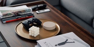 A wooden coffee table featuring a gold tray with a black game controller, a white geometric decorative object, and a stack of beige coasters. Various books and magazines are stacked nearby, along with a remote control and a smartphone.