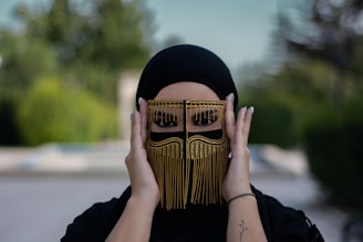 A person wearing a stylish, ornate black headscarf and a decorative face covering with fringe and gold embellishments, framed against a blurred natural background.