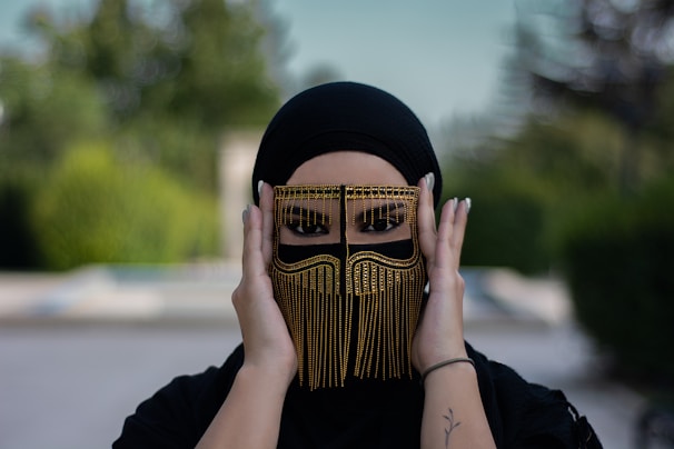 A person wearing a stylish, ornate black headscarf and a decorative face covering with fringe and gold embellishments, framed against a blurred natural background.