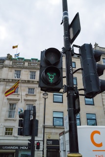 A traffic light displaying a green light in the shape of a Taurus zodiac symbol. The background consists of a stone building with several rectangular windows, a visible sign for 'Uganda House', and two flags flying. The sky appears overcast. Additional elements include a white truck partially visible on the right.