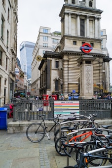 A busy urban street scene featuring a historic building with classical architecture. The entrance to a London Underground station is prominently displayed. Several bicycles are parked along the sidewalk, and people are visible in the background engaging in various activities. Modern skyscrapers can be seen in the distance, contrasting with the older architecture.