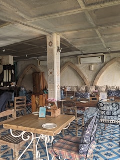 A rustic indoor café setting with wooden tables and chairs, decorative tiled flooring, and arched stone walls. The ambiance is cozy with patterned cushions and floral centerpieces on the tables. There's a mix of metal and wooden furniture, creating an eclectic and inviting atmosphere.