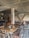 A rustic indoor café setting with wooden tables and chairs, decorative tiled flooring, and arched stone walls. The ambiance is cozy with patterned cushions and floral centerpieces on the tables. There's a mix of metal and wooden furniture, creating an eclectic and inviting atmosphere.
