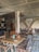 A rustic indoor café setting with wooden tables and chairs, decorative tiled flooring, and arched stone walls. The ambiance is cozy with patterned cushions and floral centerpieces on the tables. There's a mix of metal and wooden furniture, creating an eclectic and inviting atmosphere.