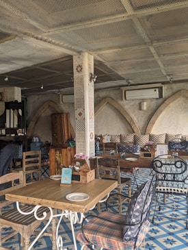 A rustic indoor caf&eacute; setting with wooden tables and chairs, decorative tiled flooring, and arched stone walls. The ambiance is cozy with patterned cushions and floral centerpieces on the tables. There's a mix of metal and wooden furniture, creating an eclectic and inviting atmosphere.